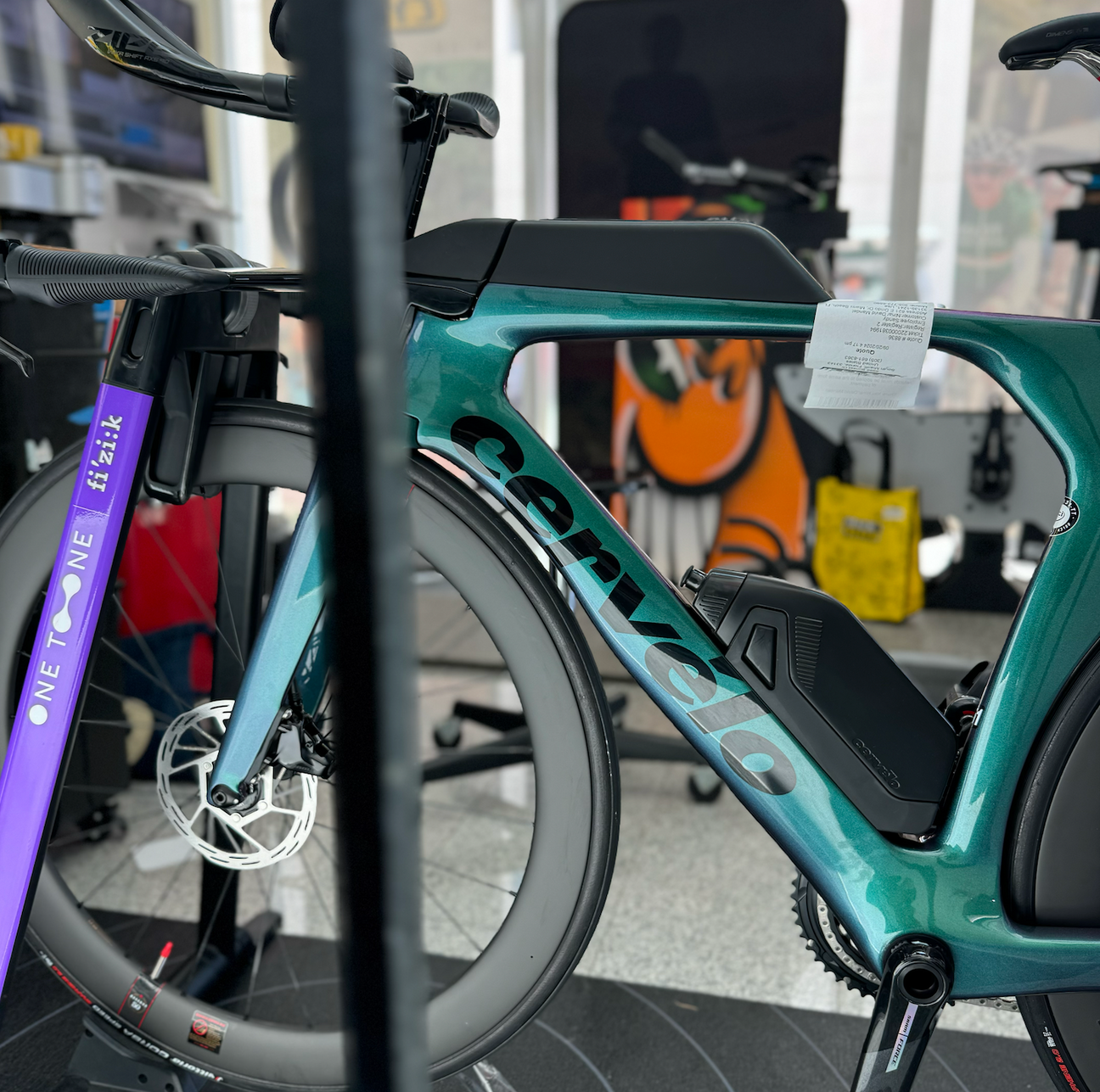 A Cervelo P5 SRAM Force eTap AXS Bike in Twighlight Chameleon color mounted on a wahoo kickr rollr. The bike appears green inside the mack cycle fit studio. Behind the bike is an orange atomik grafiti painting smiling through the bike frame